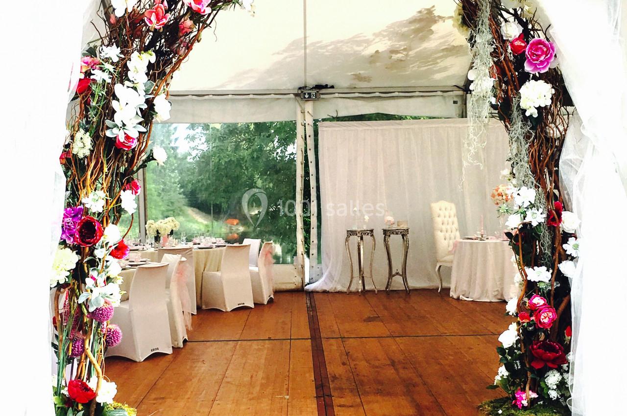 Arche décorée de fleurs colorées dans une tente de réception avec tables et chaises en arrière-plan.