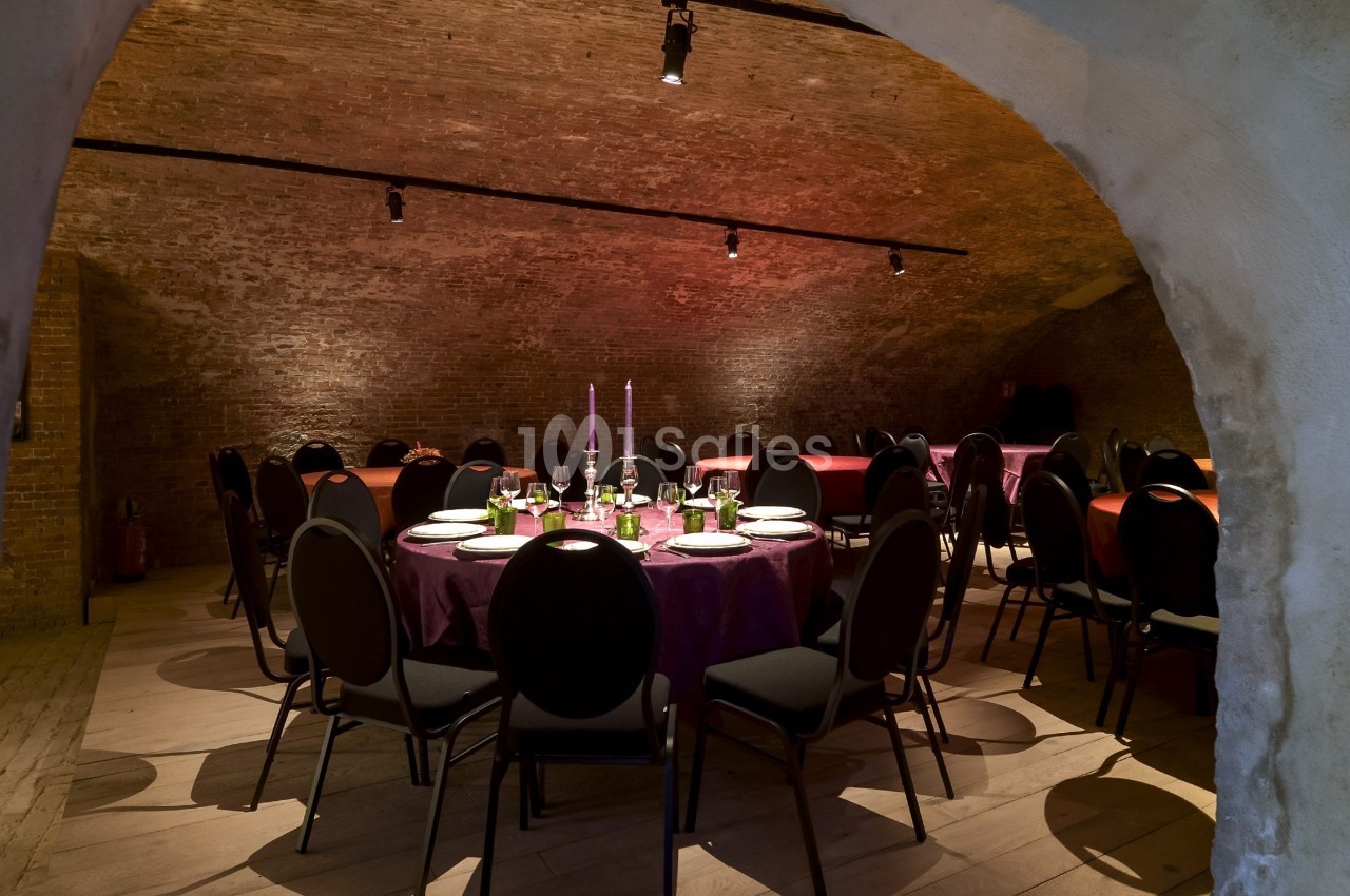 Salle voûtée avec des tables rondes dressées pour un repas, éclairée par des bougies et des lumières tamisées.