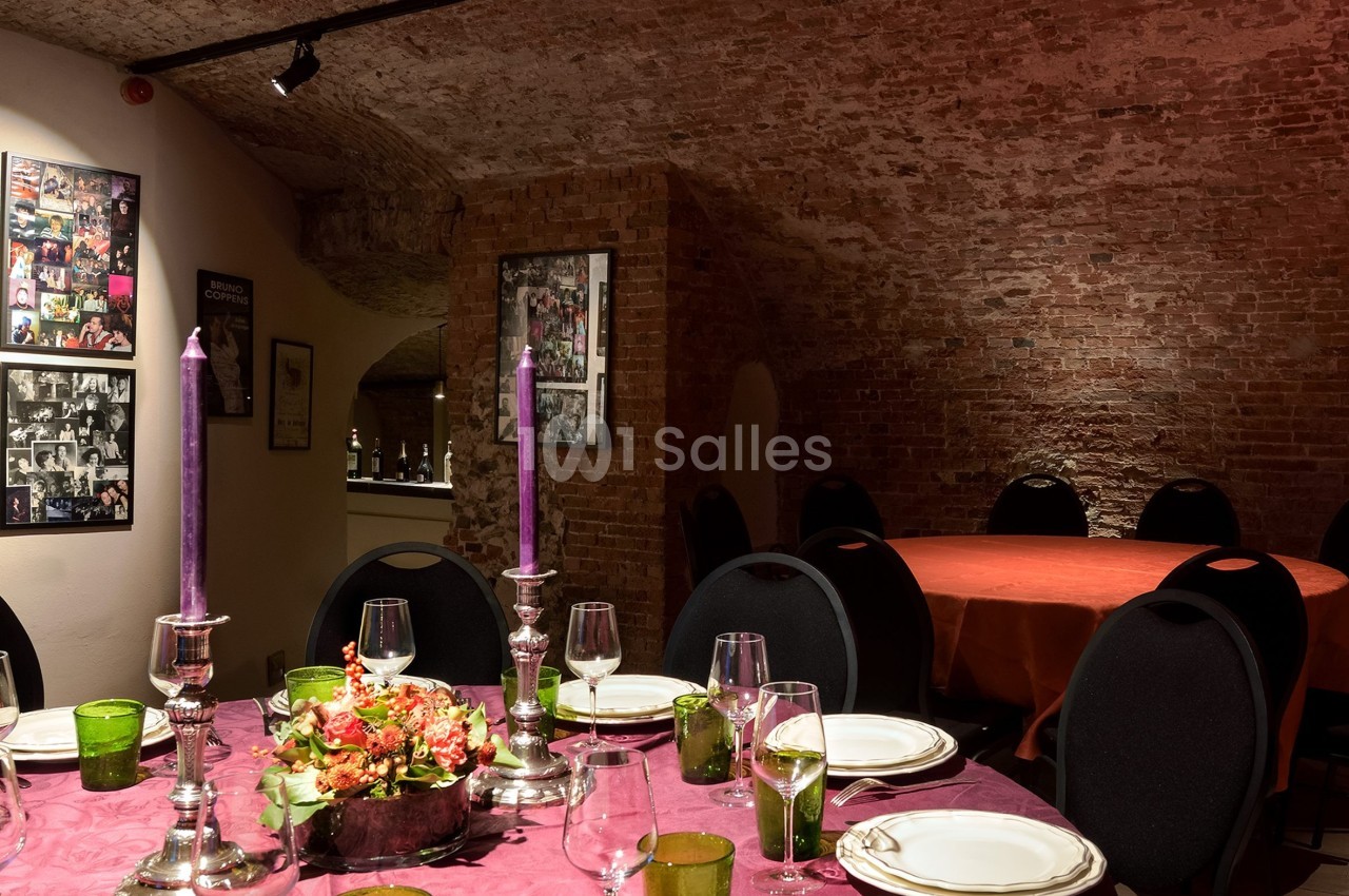 Salle voûtée en briques avec tables dressées, nappes colorées, chandeliers et décoration florale.