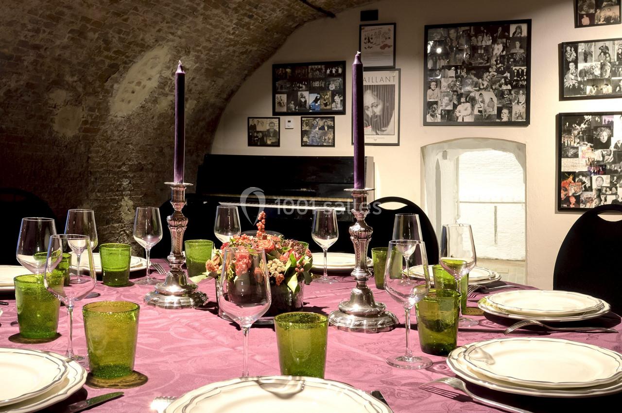 Location salle Bruxelles (Bruxelles-Capitale) - Théâtre de la Samaritaine #4 Table dressée avec nappe violette, vaisselle élégante, bougies allumées, dans une salle voûtée décorée de photos.