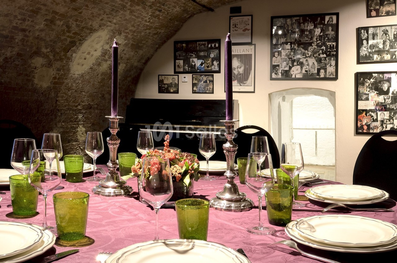 Table dressée avec nappe violette, vaisselle élégante, bougies allumées, dans une salle voûtée décorée de photos.