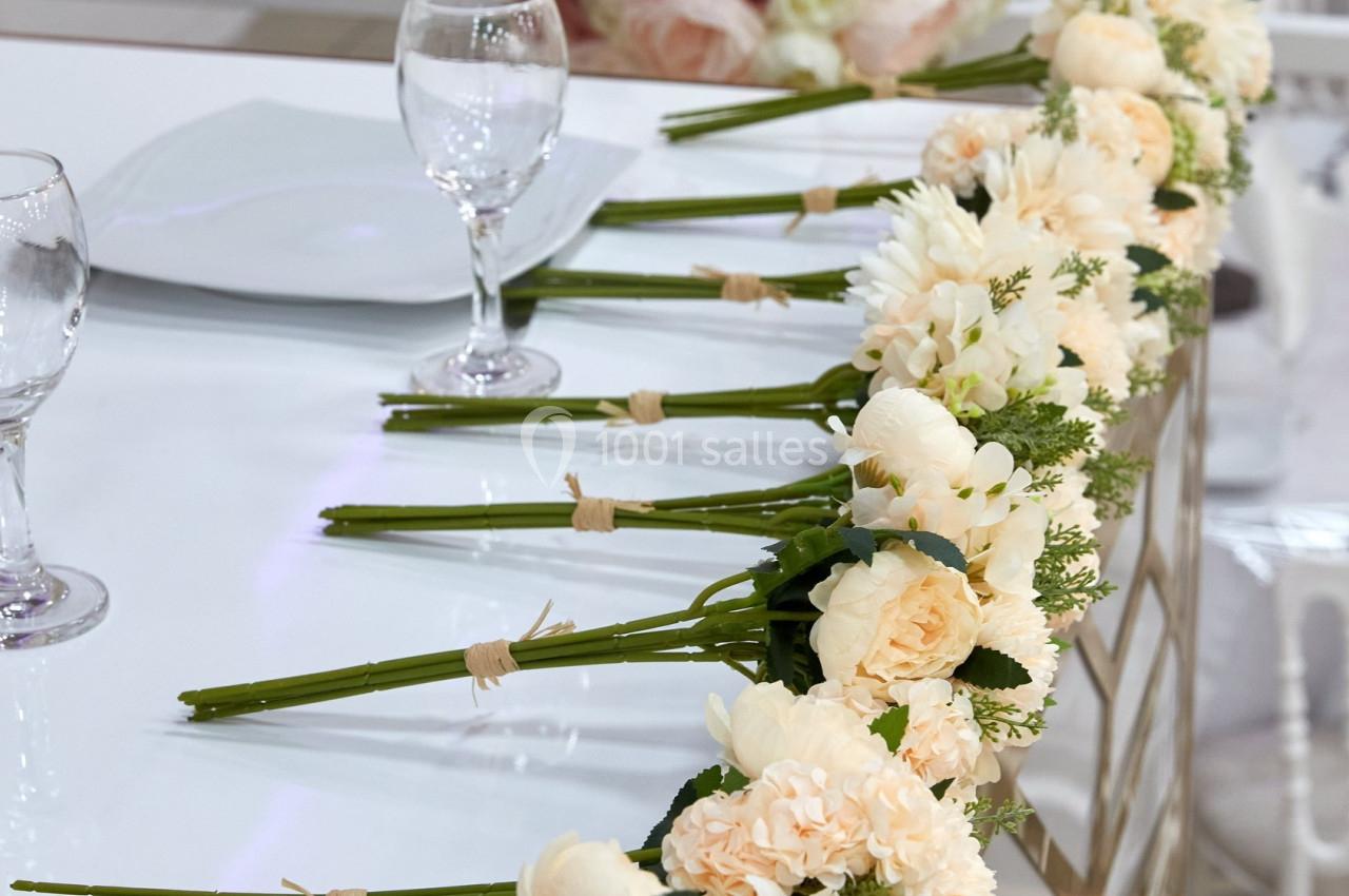 Bouquets de fleurs crème alignés sur une table décorative, avec un centre de table composé de roses pastel en arrière-plan.