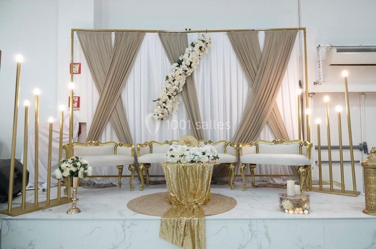 Décor de cérémonie avec rideaux beige et blanc, fleurs blanches, chandeliers dorés et table ornée de tissu doré.