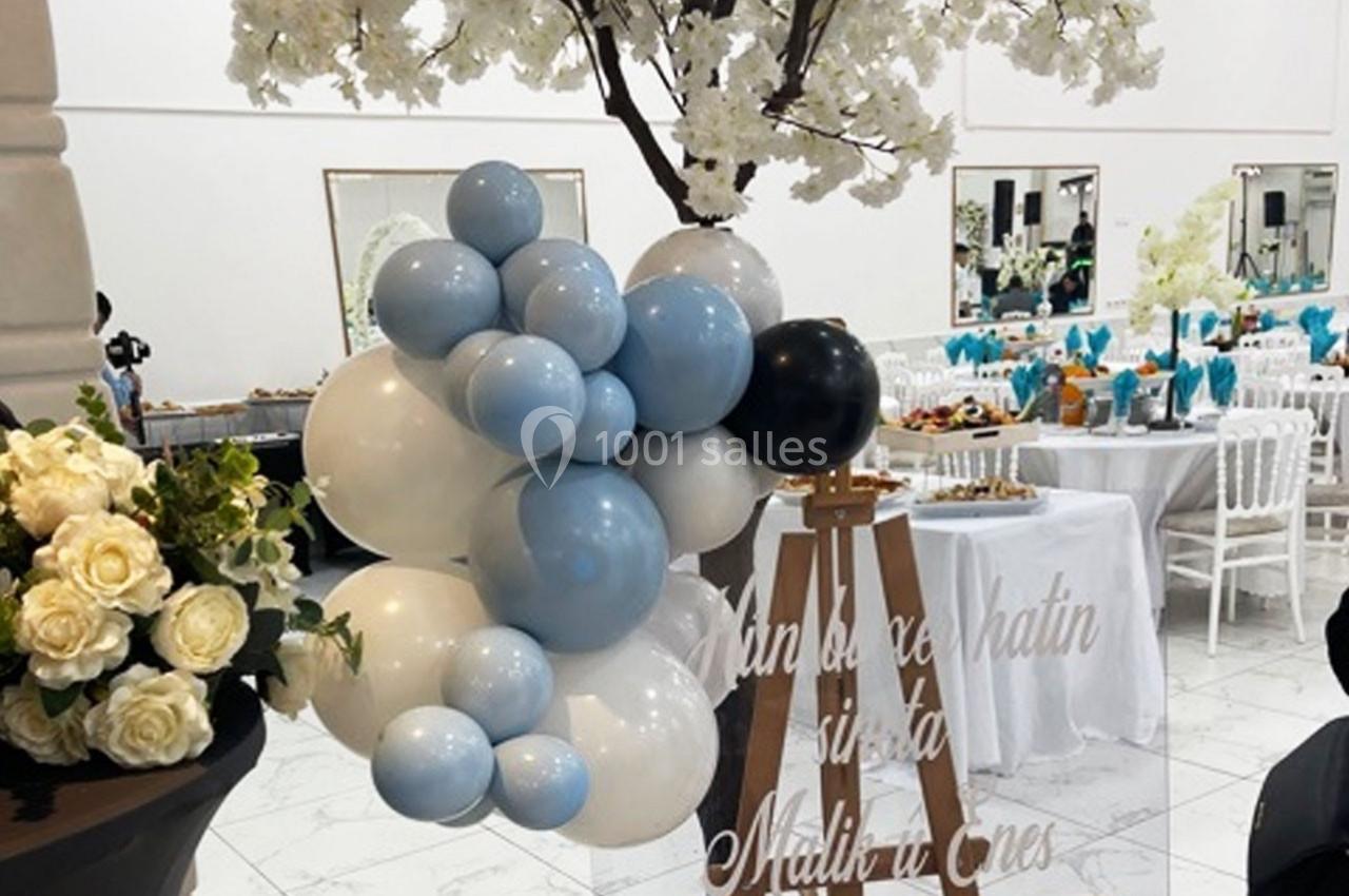 Décoration de fête avec des ballons bleus, blancs et noirs, un chevalet et des fleurs blanches dans une salle élégante.