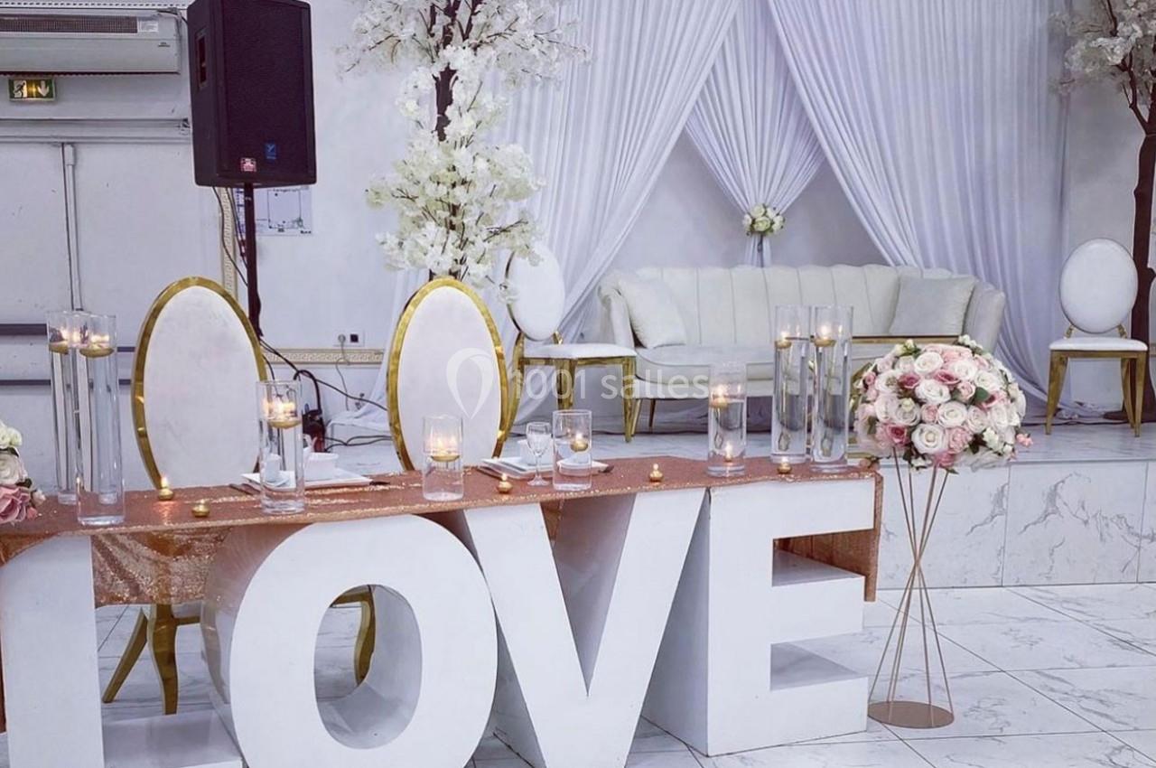 Décoration de salle de réception avec grandes lettres ’LOVE’, table ornée de bougies et fleurs, canapé et rideaux blancs.