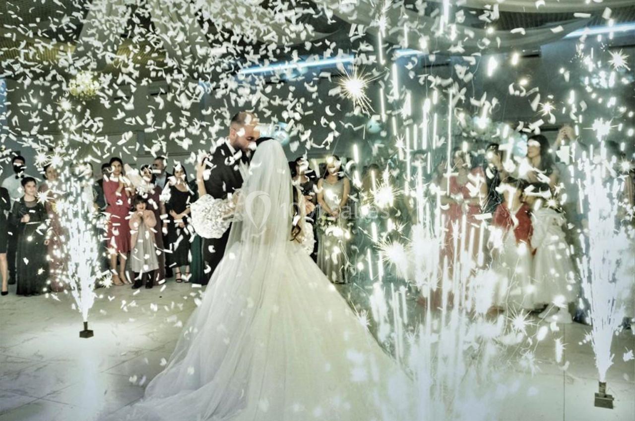 Un couple de mariés danse au centre d'une salle illuminée par des étincelles et des confettis, entouré d'invités.