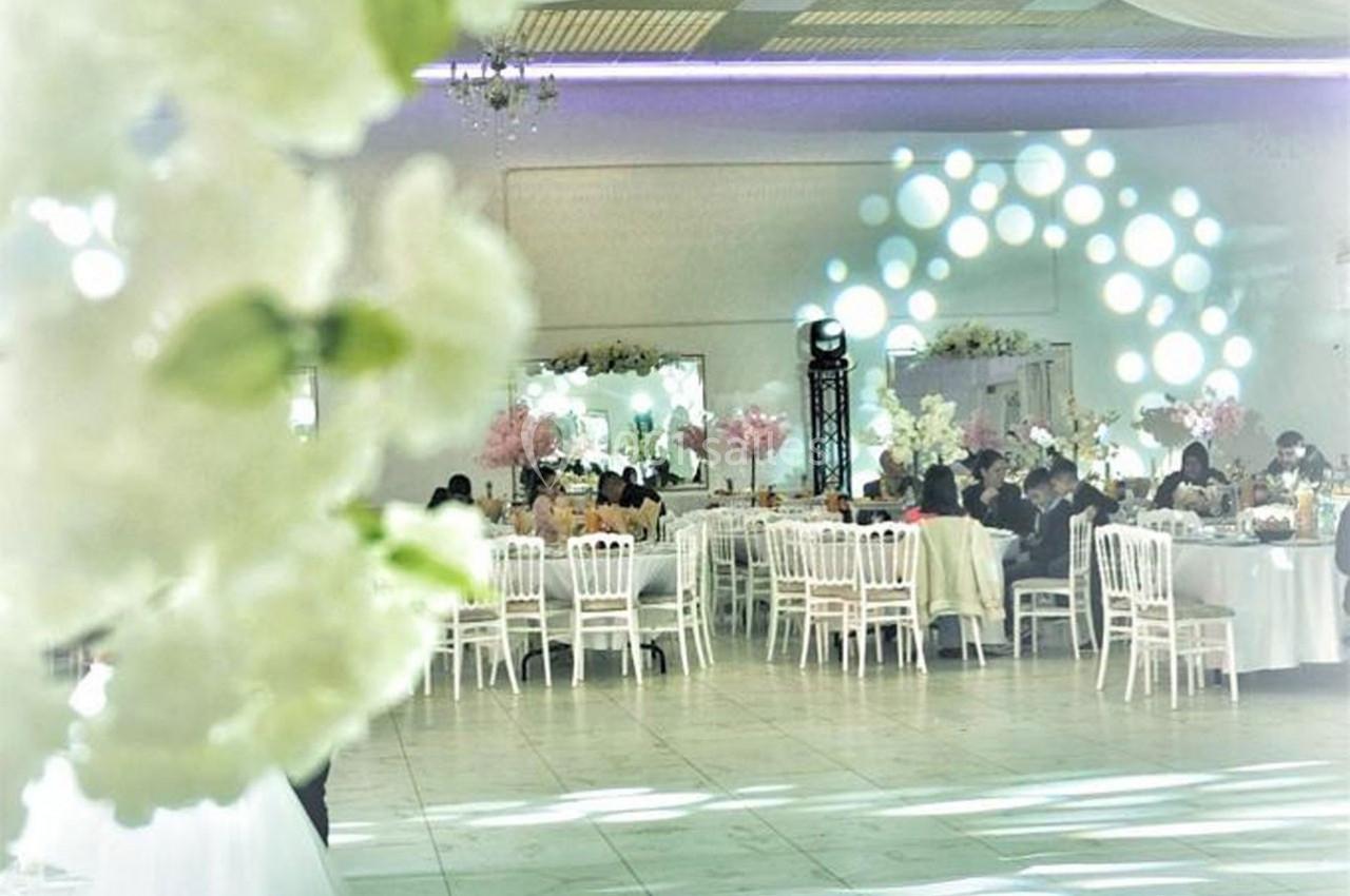 Salle de réception décorée avec des fleurs blanches et roses, tables dressées et invités assis lors d'un événement.