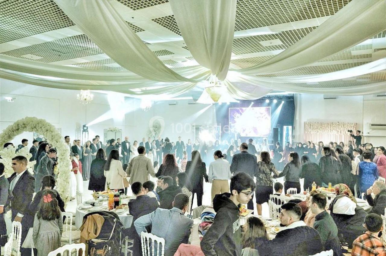 Salle de réception décorée avec des drapés blancs, accueillant un grand rassemblement de personnes autour d'une piste de…