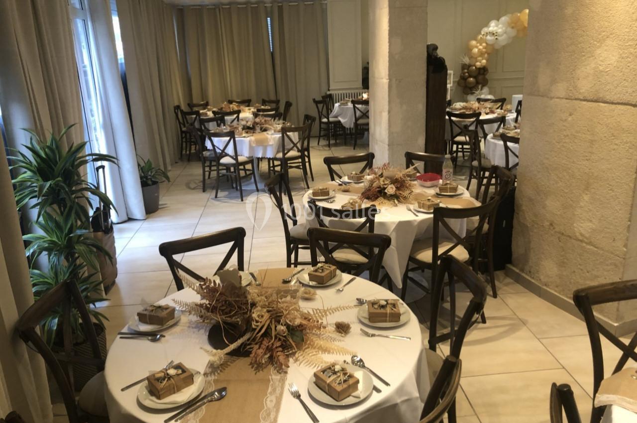 Salle décorée pour un événement, avec des tables rondes dressées, ornées de centres de table naturels et cadeaux.