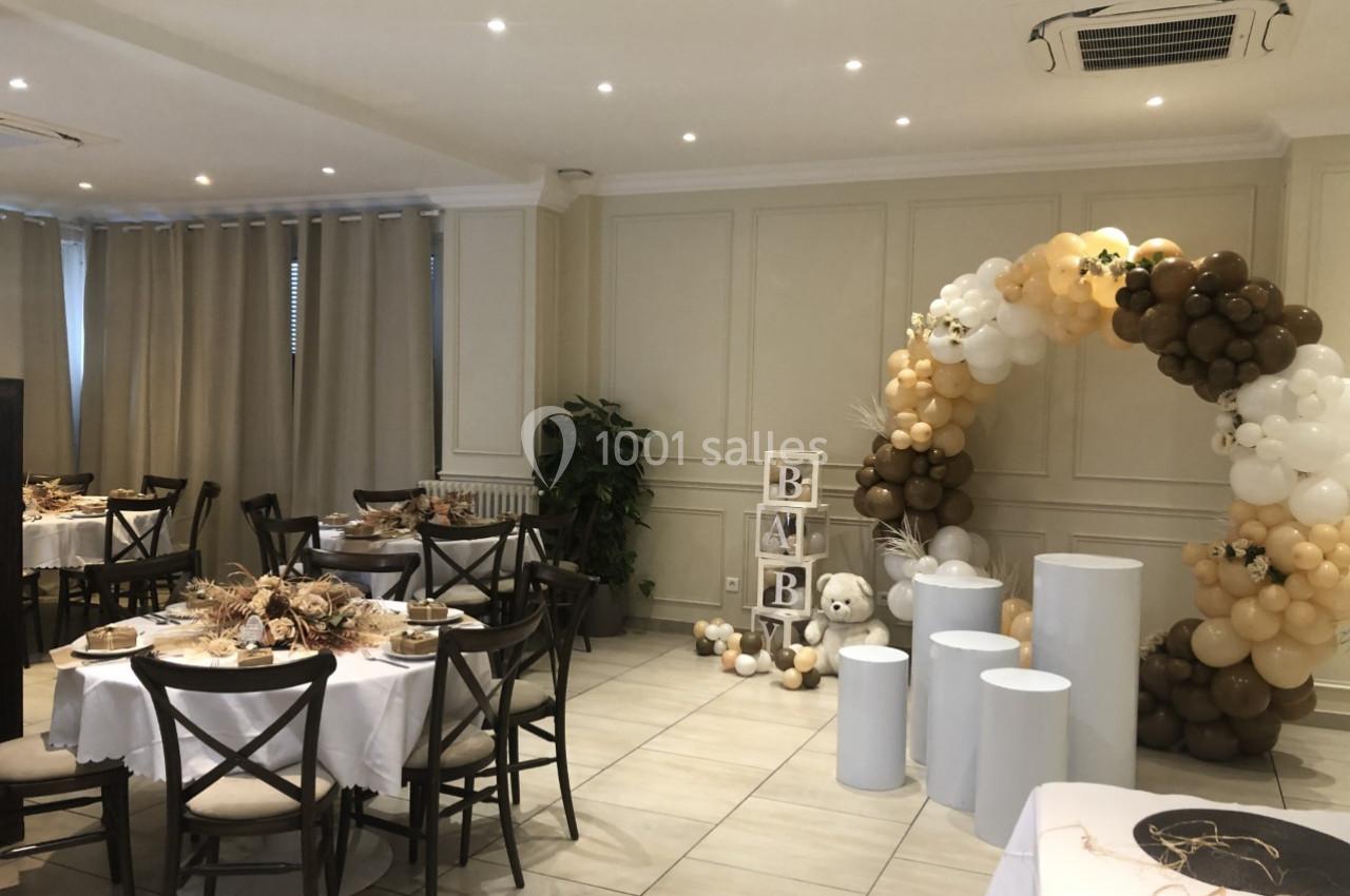 Salle décorée pour une fête avec arche de ballons beige et blanc, tables dressées et cubes formant le mot ’BABY’.