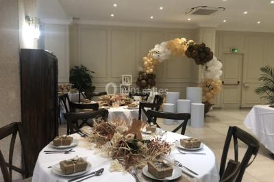 Salle décorée pour un événement, avec des tables rondes dressées, ornées de centres de table naturels et cadeaux.