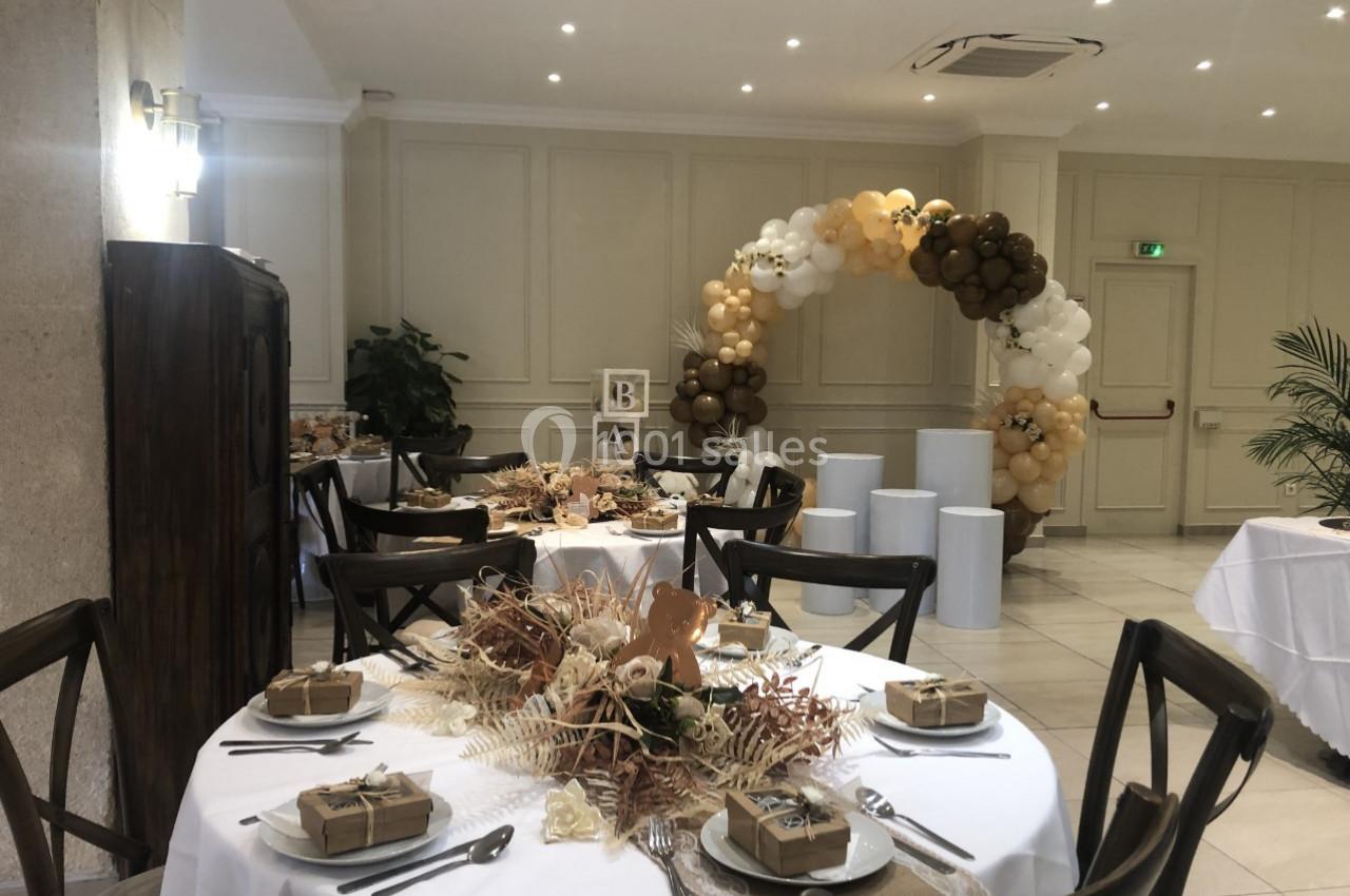 Salle décorée pour un événement avec tables dressées, centre de table floral et arche de ballons beige et marron.