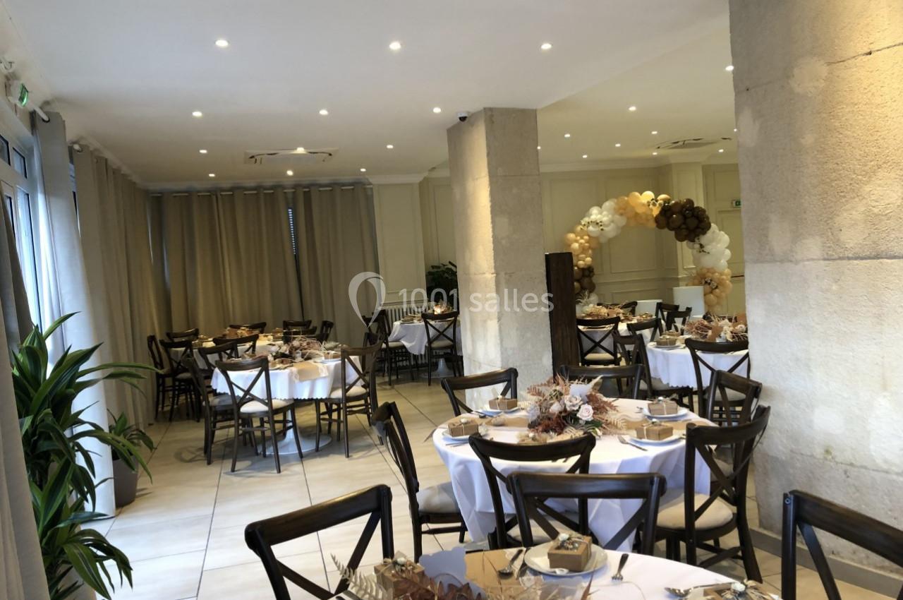 Salle de réception décorée pour un événement, avec tables rondes dressées, chaises en bois et arche florale en arrière-plan.