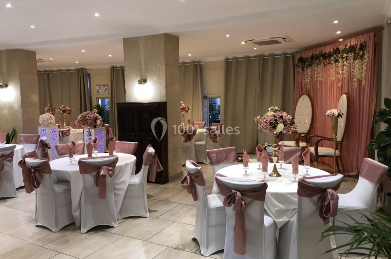 Salle de réception décorée avec des tables rondes, nappes blanches, chaises ornées de rubans roses et arrière-plan fleuri.