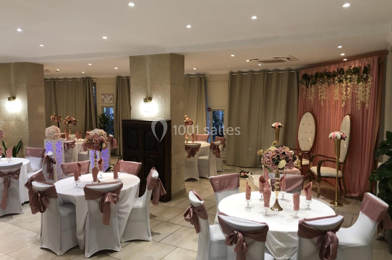 Salle de réception décorée avec des tables rondes, nappes blanches, chaises ornées de rubans roses et un fond fleuri.