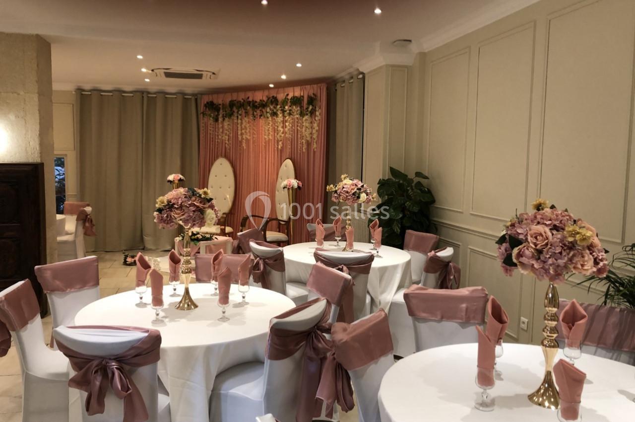 Salle décorée pour un événement avec tables rondes, nappes blanches, chaises ornées de nœuds roses et fleurs en centre de…