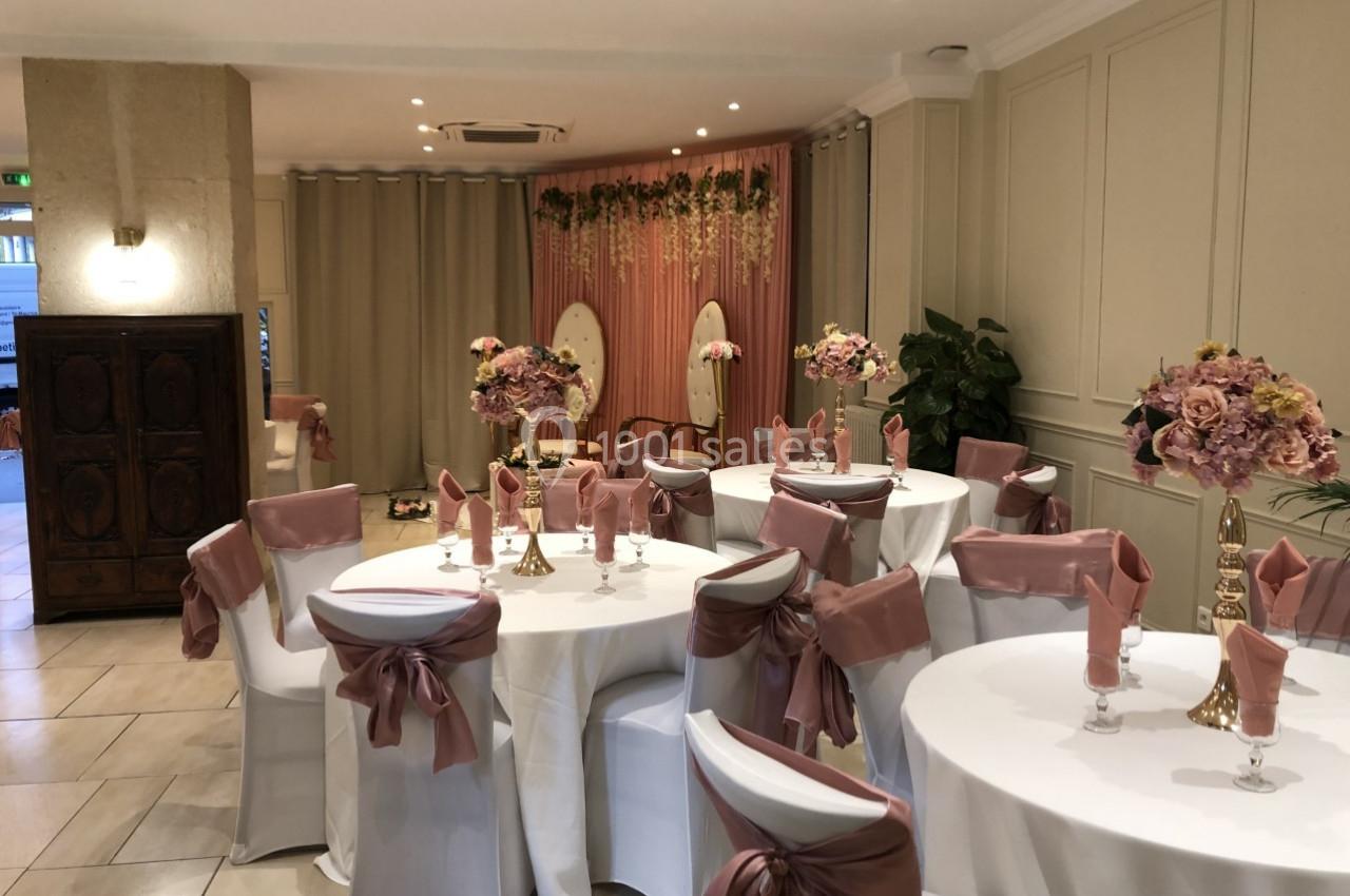 Salle décorée pour un événement avec des tables rondes, nappes blanches, chaises ornées de nœuds roses et fleurs en centre…