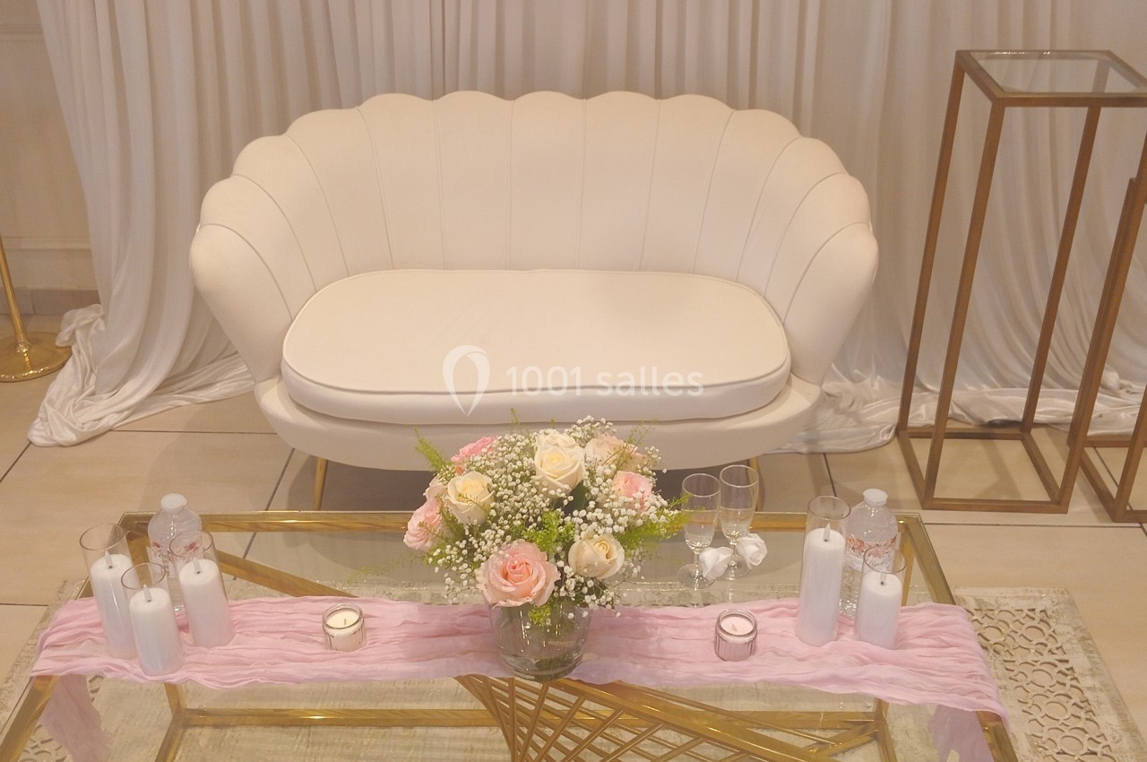 Canapé blanc devant un rideau, table basse dorée décorée de bougies, fleurs et tissu rose.