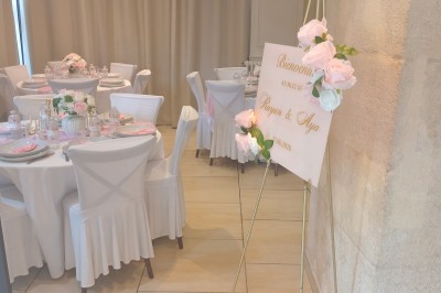 Table ronde élégamment dressée avec nappes blanches, serviettes roses pliées, vaisselle et bouquet de fleurs au centre.