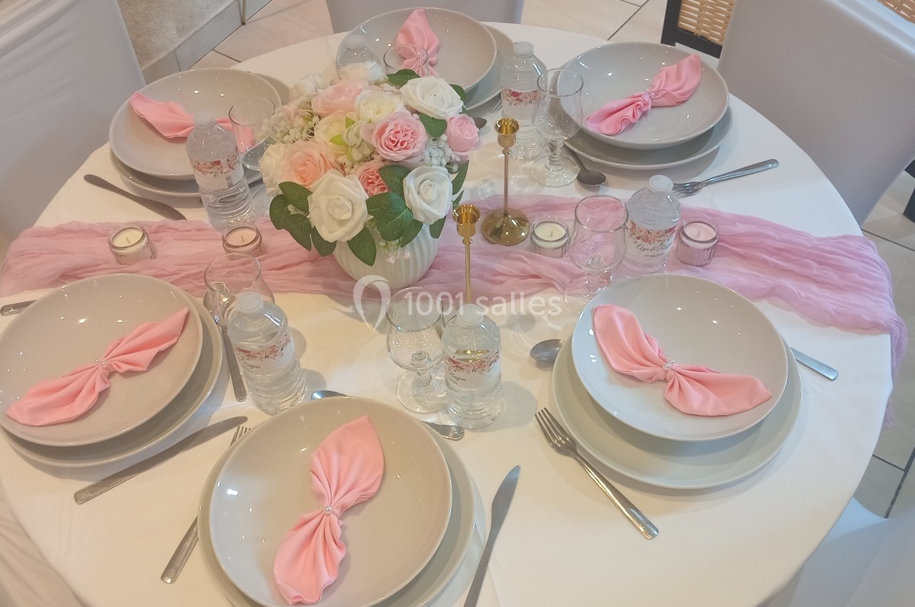 Table ronde élégamment dressée avec nappes blanches, serviettes roses, vaisselle blanche et centre de table floral.