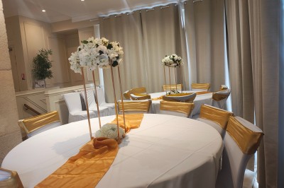 Table ronde élégamment dressée avec nappes blanches, serviettes roses pliées, vaisselle et bouquet de fleurs au centre.