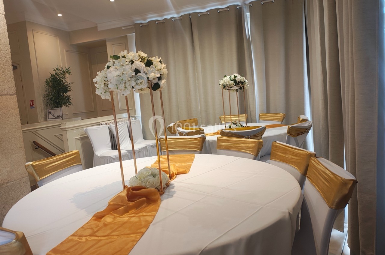 Salle décorée pour un événement avec tables rondes, nappes blanches, chemins de table orange et centres de table fleuris.