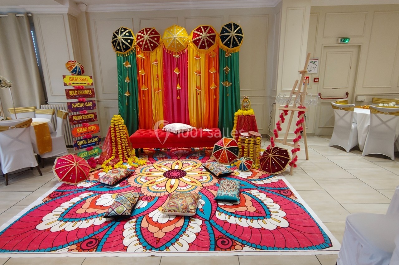 Décor coloré avec tapis floral, coussins, parasols et guirlandes, installé dans une salle de réception lumineuse.