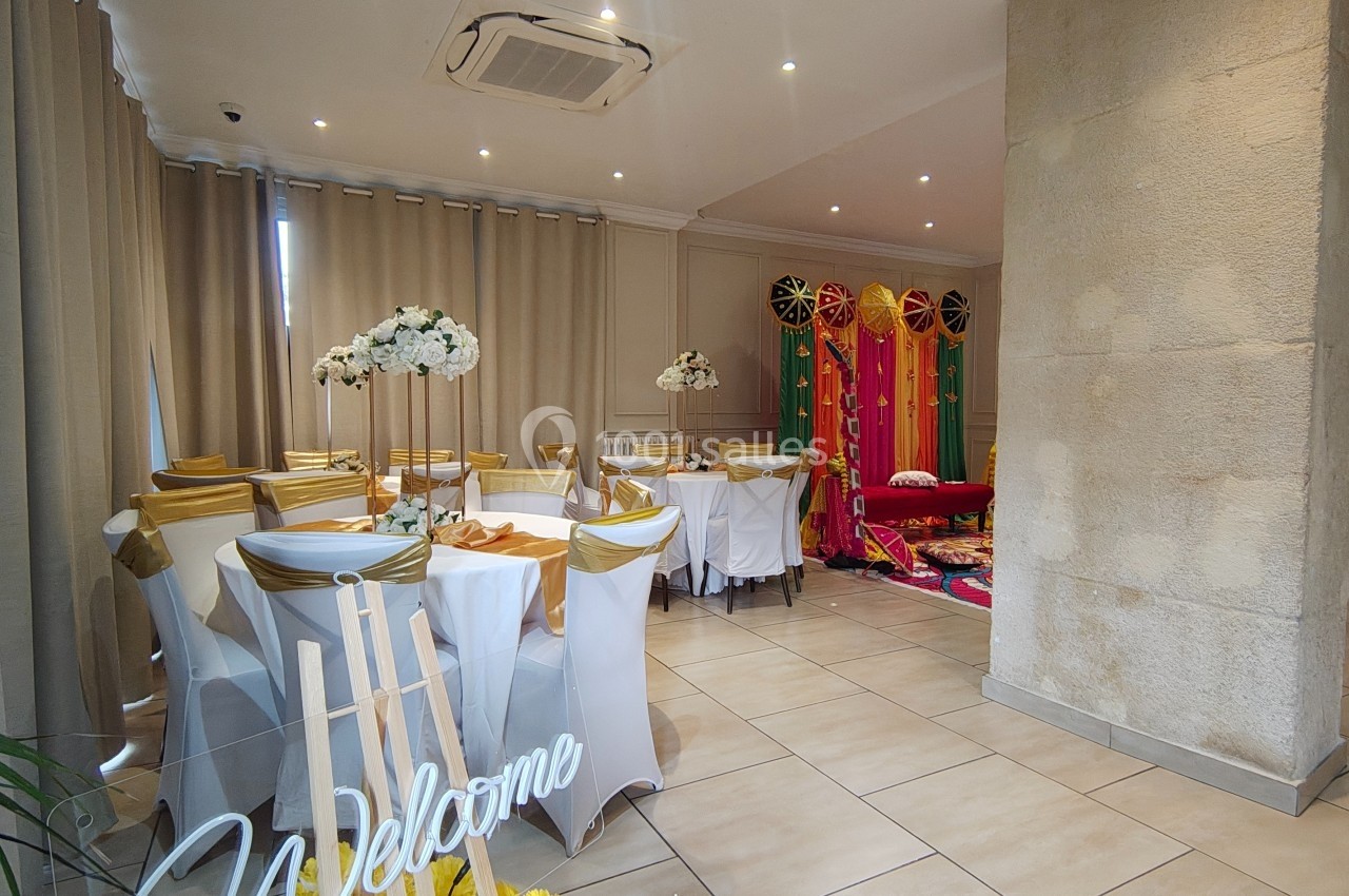 Salle décorée pour un événement, avec chaises blanches ornées de nœuds dorés, fleurs et rideaux colorés en arrière-plan.