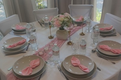 Table ronde élégamment dressée avec nappes blanches, serviettes roses pliées, vaisselle et bouquet de fleurs au centre.