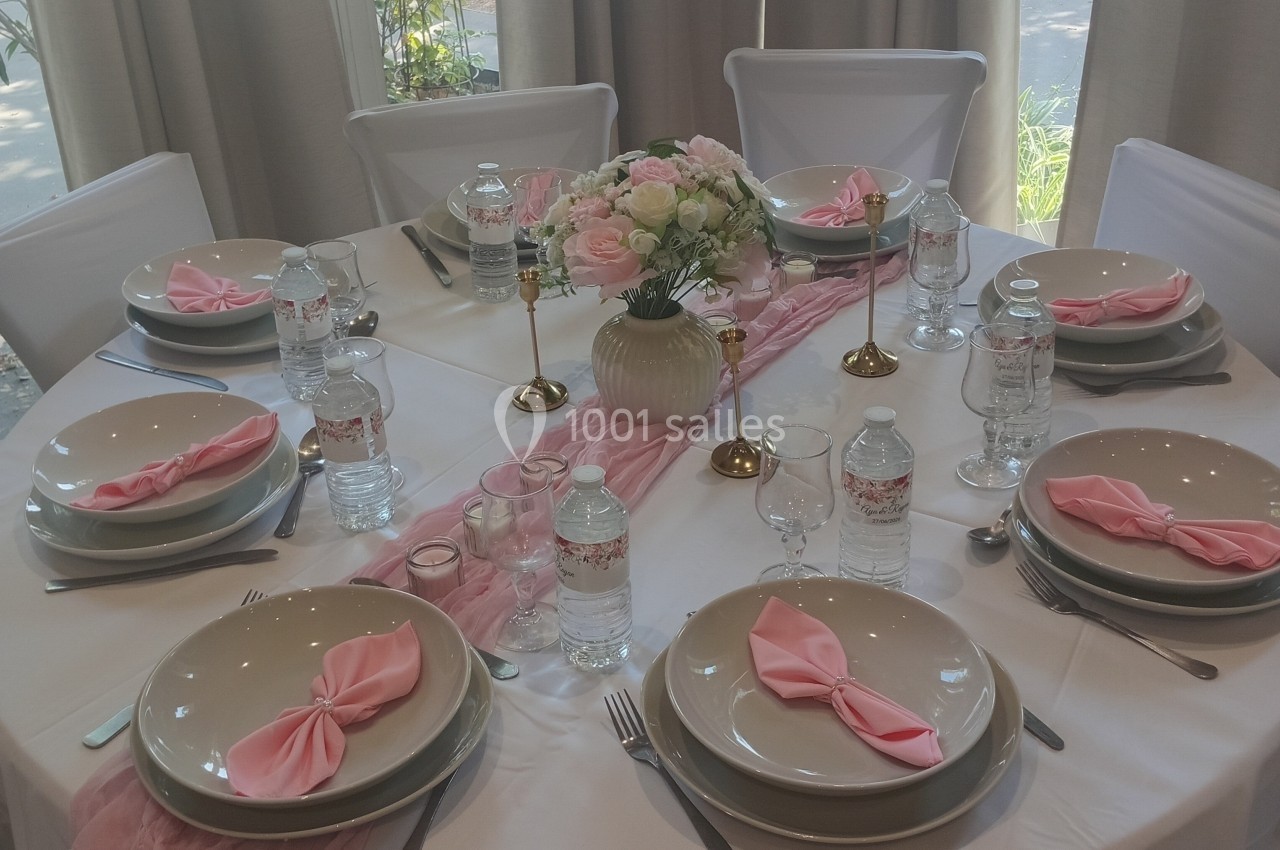 Table élégamment dressée avec vaisselle beige, serviettes roses pliées, bougies dorées et centre de table floral.
