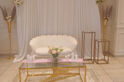 Table ronde élégamment dressée avec nappes blanches, serviettes roses pliées, vaisselle et bouquet de fleurs au centre.