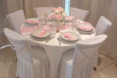 Table ronde élégamment dressée avec nappes blanches, serviettes roses pliées, vaisselle et bouquet de fleurs au centre.