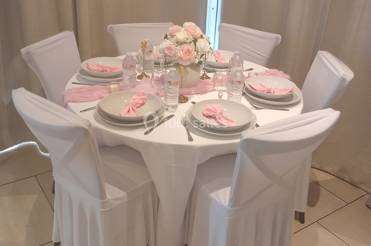 Table ronde élégamment dressée avec nappes blanches, serviettes roses pliées, vaisselle et bouquet de fleurs au centre.