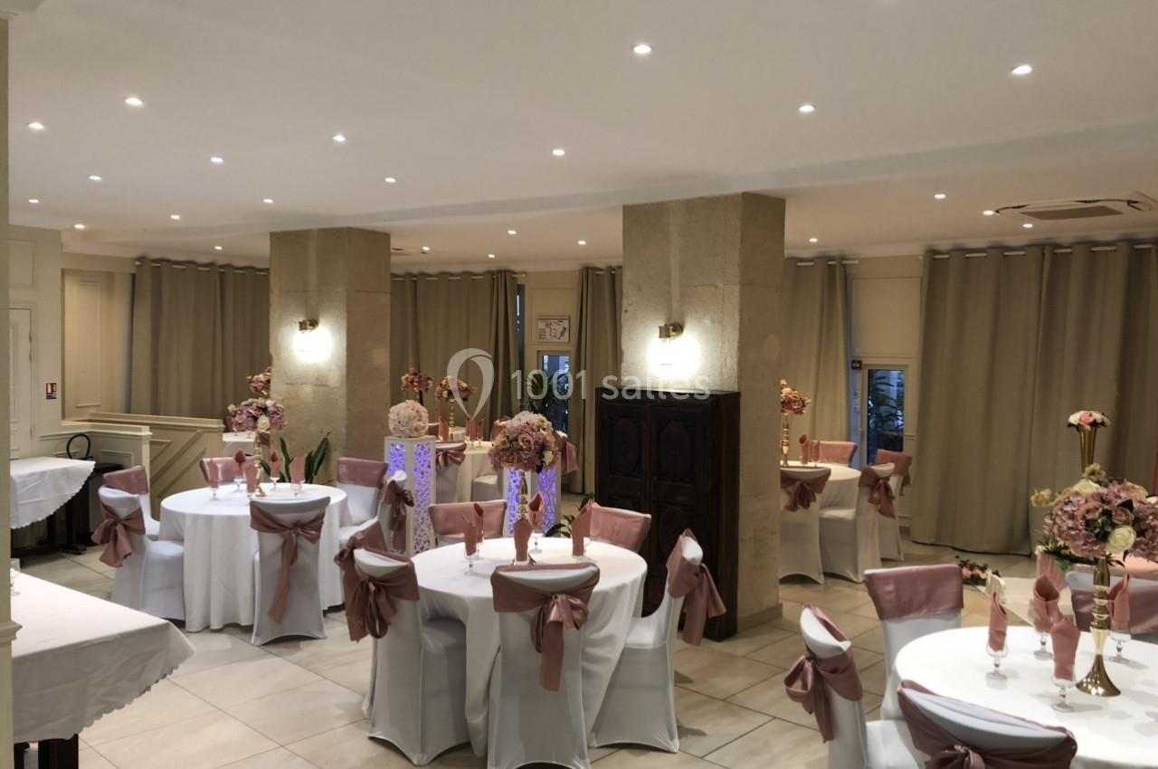 Salle de réception décorée avec des tables rondes, nappes blanches, chaises ornées de nœuds roses et centres de table…