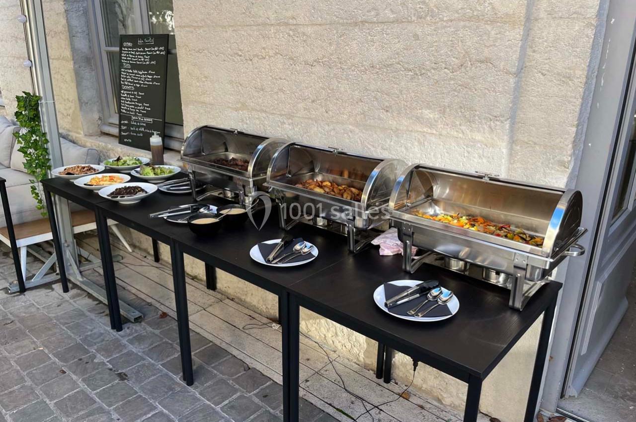 Buffet extérieur avec plats chauds en chafing dishes, accompagnés de salades et assiettes sur une table noire.