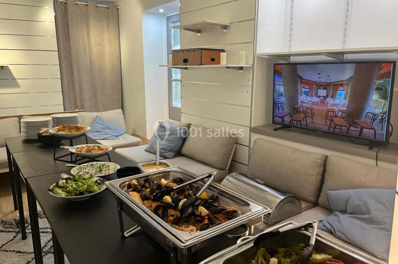 Buffet avec plats variés, salades et légumes, disposé dans un salon avec canapé, table et télévision allumée.