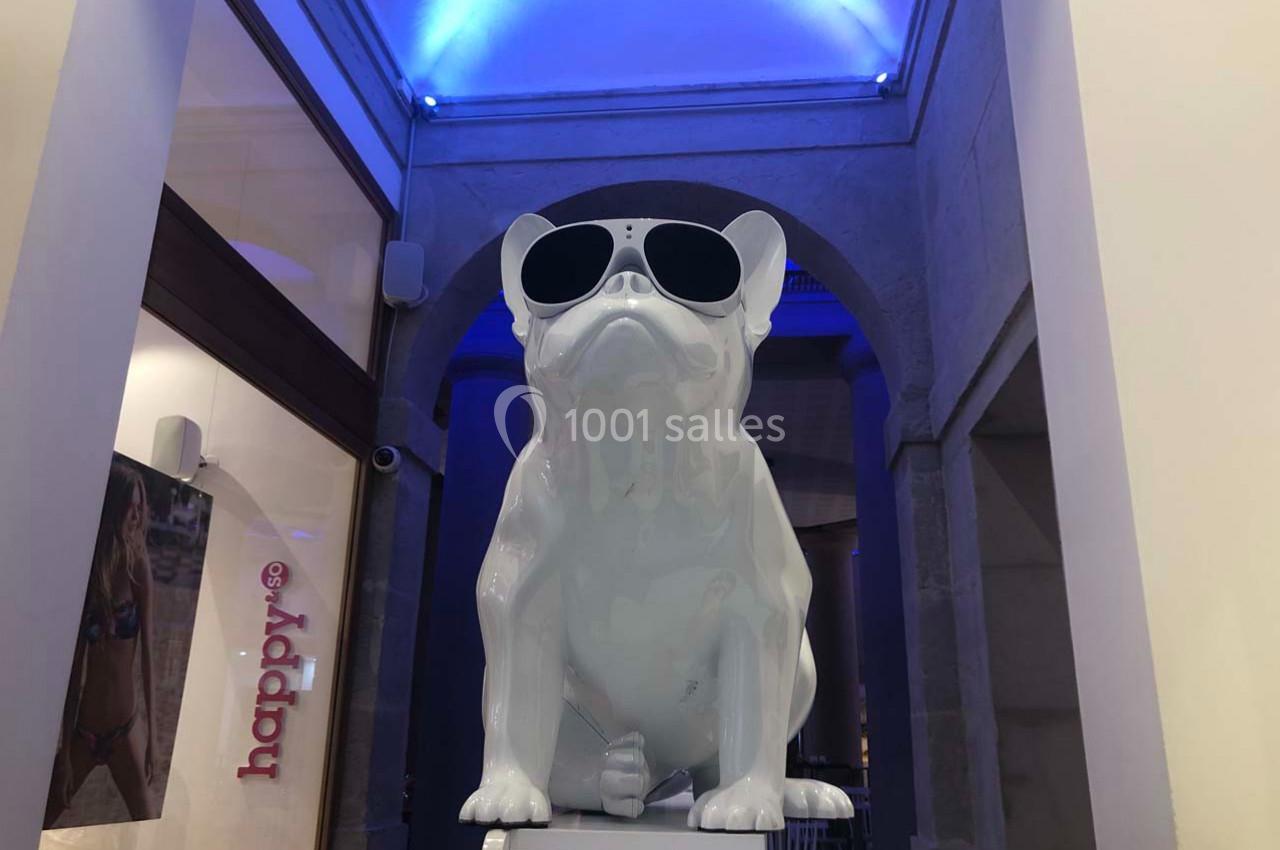 Statue blanche d'un bouledogue portant des lunettes de soleil, exposée dans un espace intérieur avec éclairage bleu.