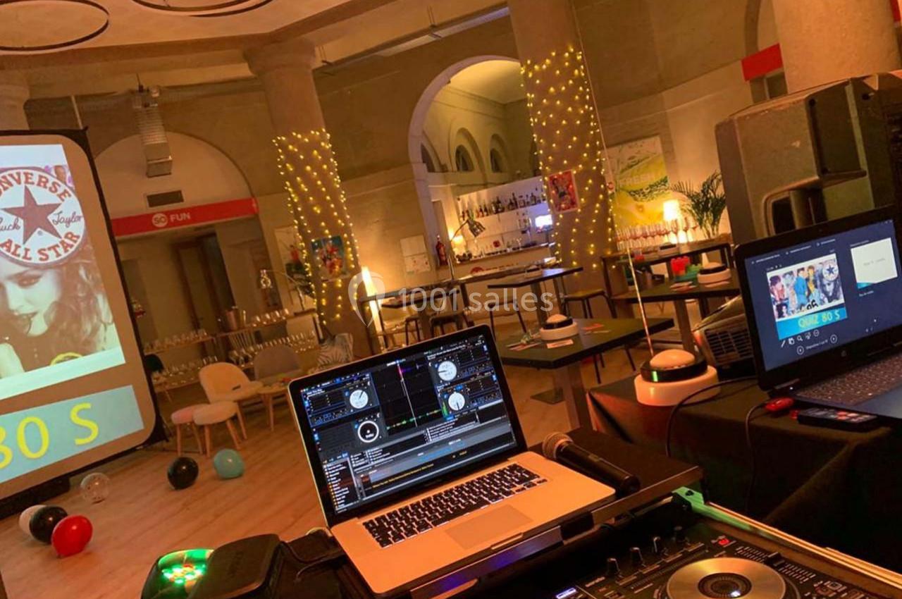 Salle décorée pour un événement avec tables, guirlandes lumineuses et matériel de DJ en premier plan.