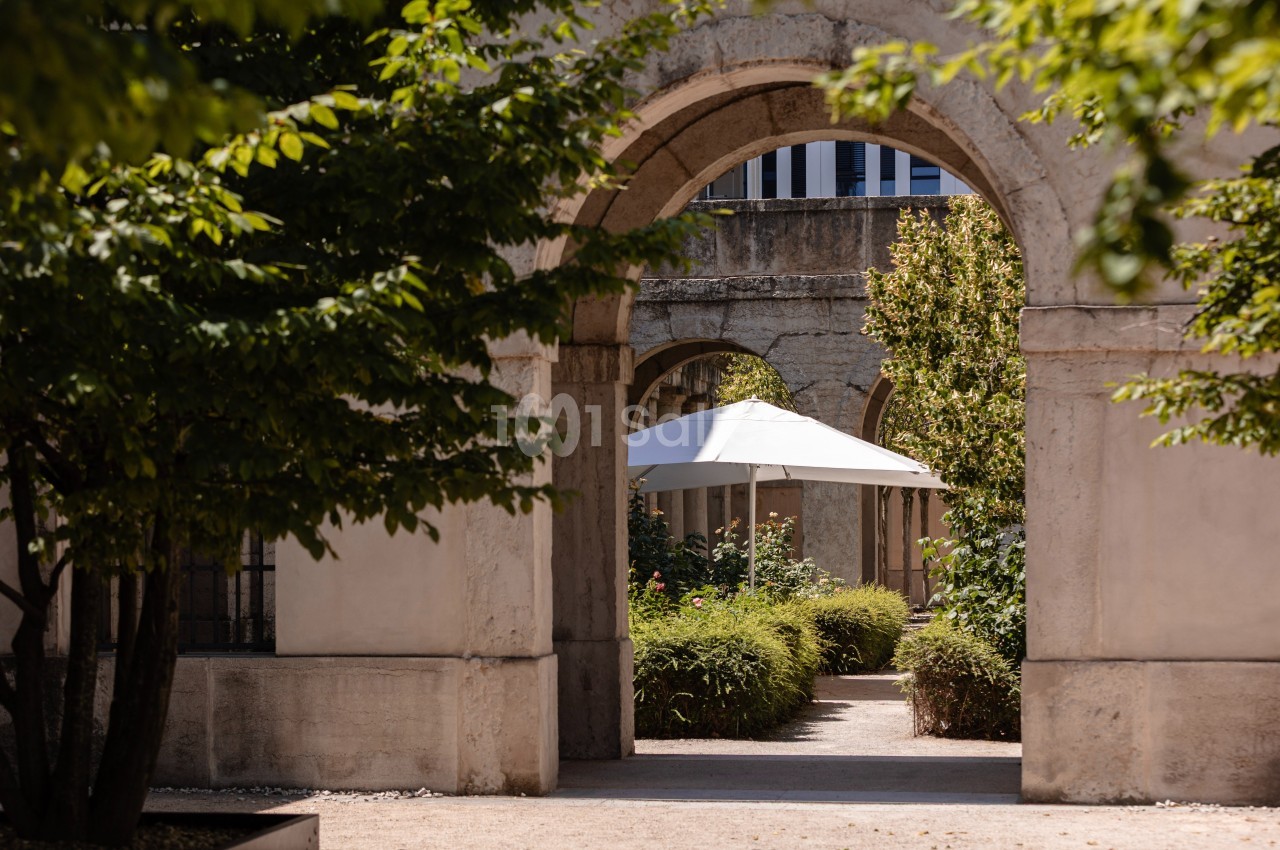 Vue d'une arche en pierre donnant sur un jardin avec des plantes, un chemin et un parasol blanc.