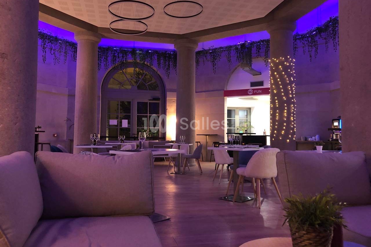Salle de restaurant élégante avec éclairage tamisé violet, colonnes, plantes suspendues et mobilier moderne.