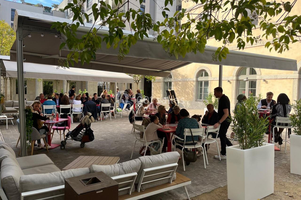 Terrasse extérieure d'un café avec des tables occupées par des clients, entourée de végétation et d'un bâtiment clair.