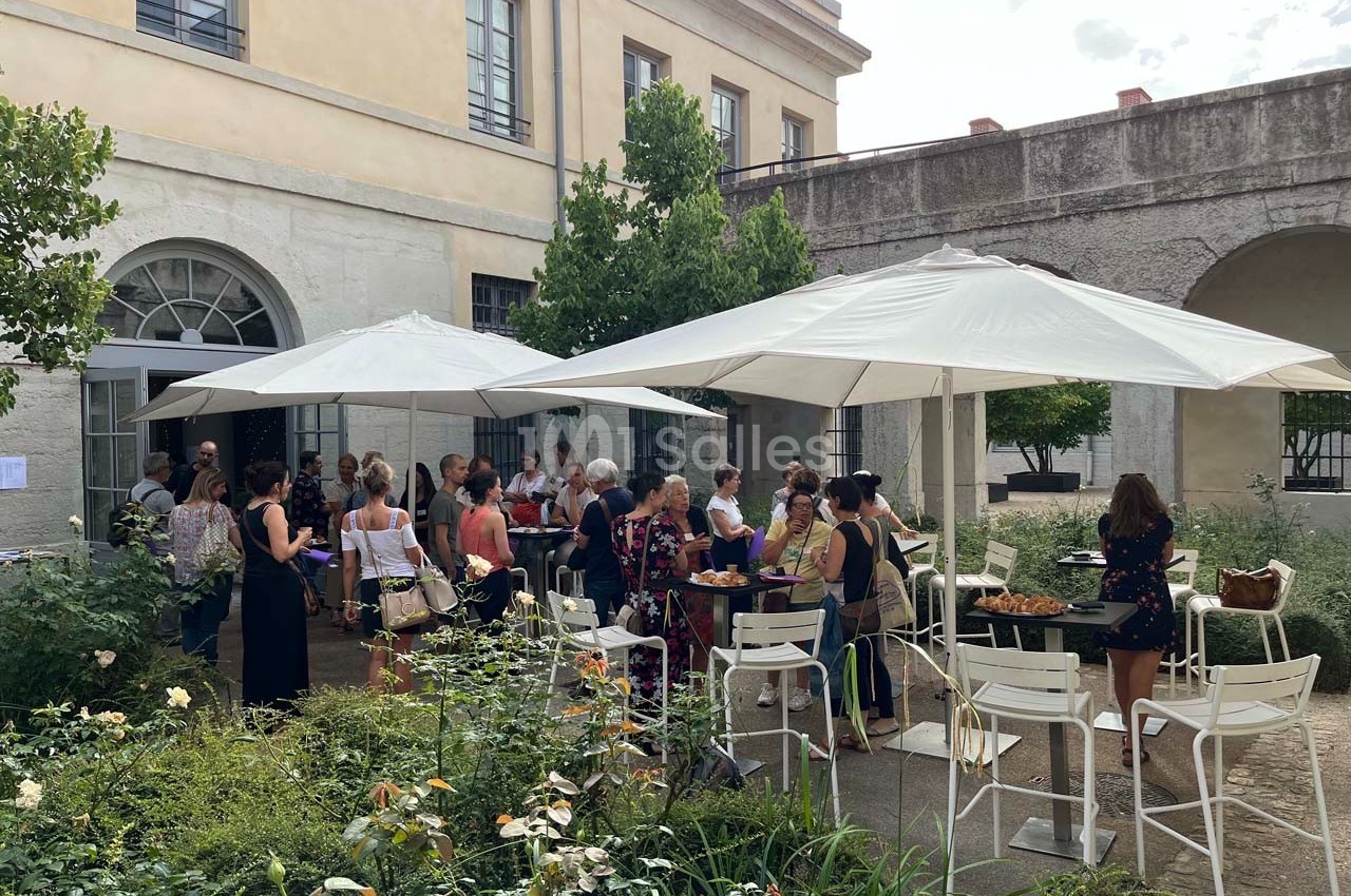 Groupe de personnes rassemblées dans une cour extérieure, discutant autour de tables sous des parasols blancs.