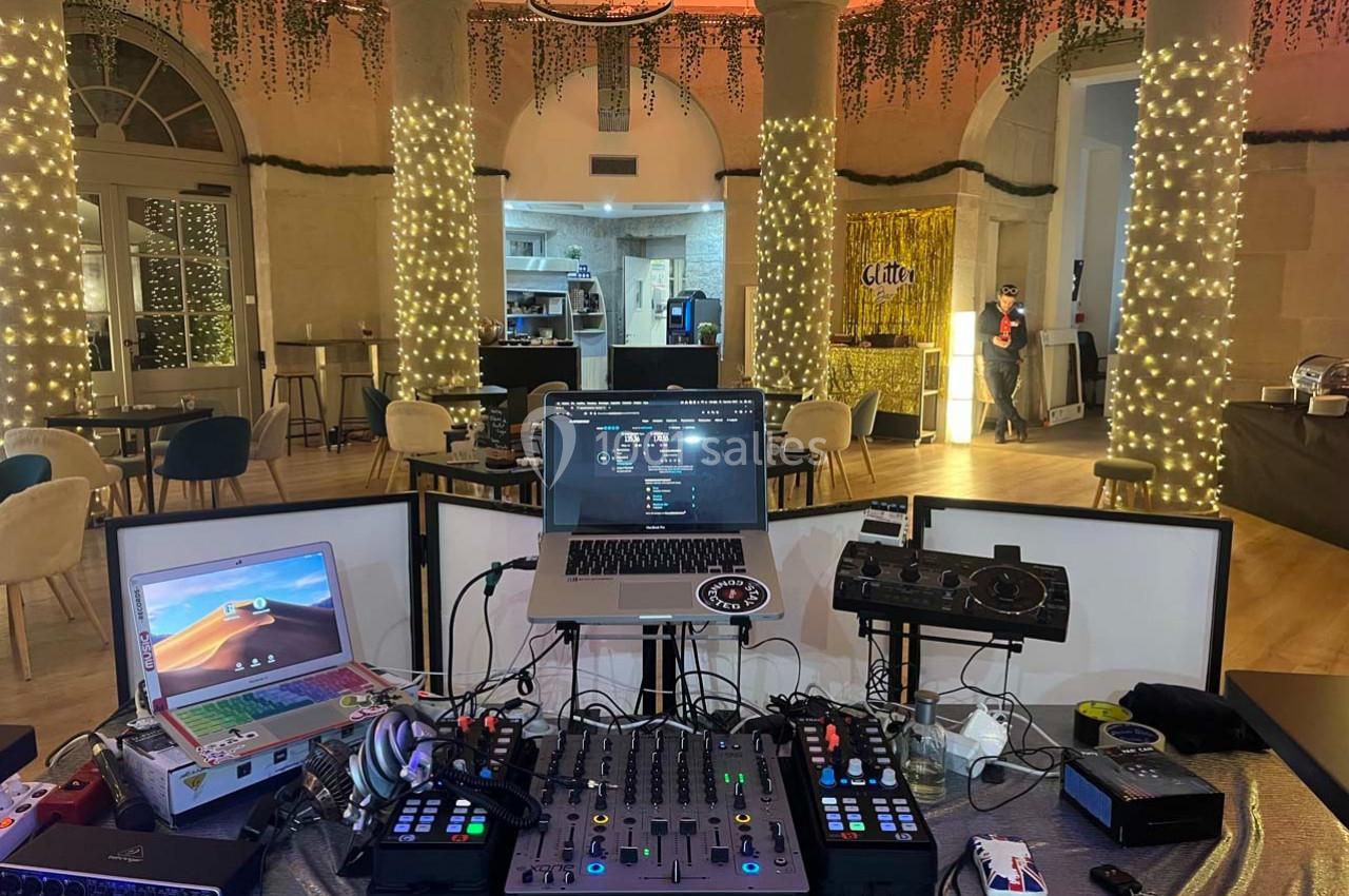 Poste de DJ avec table de mixage, ordinateurs et platines dans une salle décorée de guirlandes lumineuses.