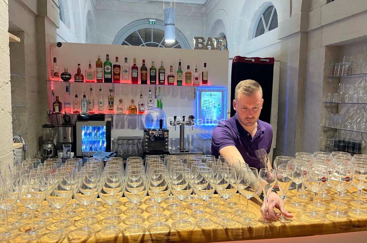Un barman en chemise violette prépare un comptoir rempli de verres à pied devant un bar éclairé.