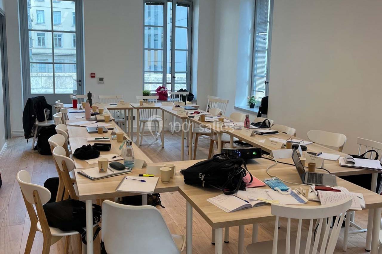 Salle de réunion lumineuse avec plusieurs tables en bois, chaises blanches et documents dispersés.