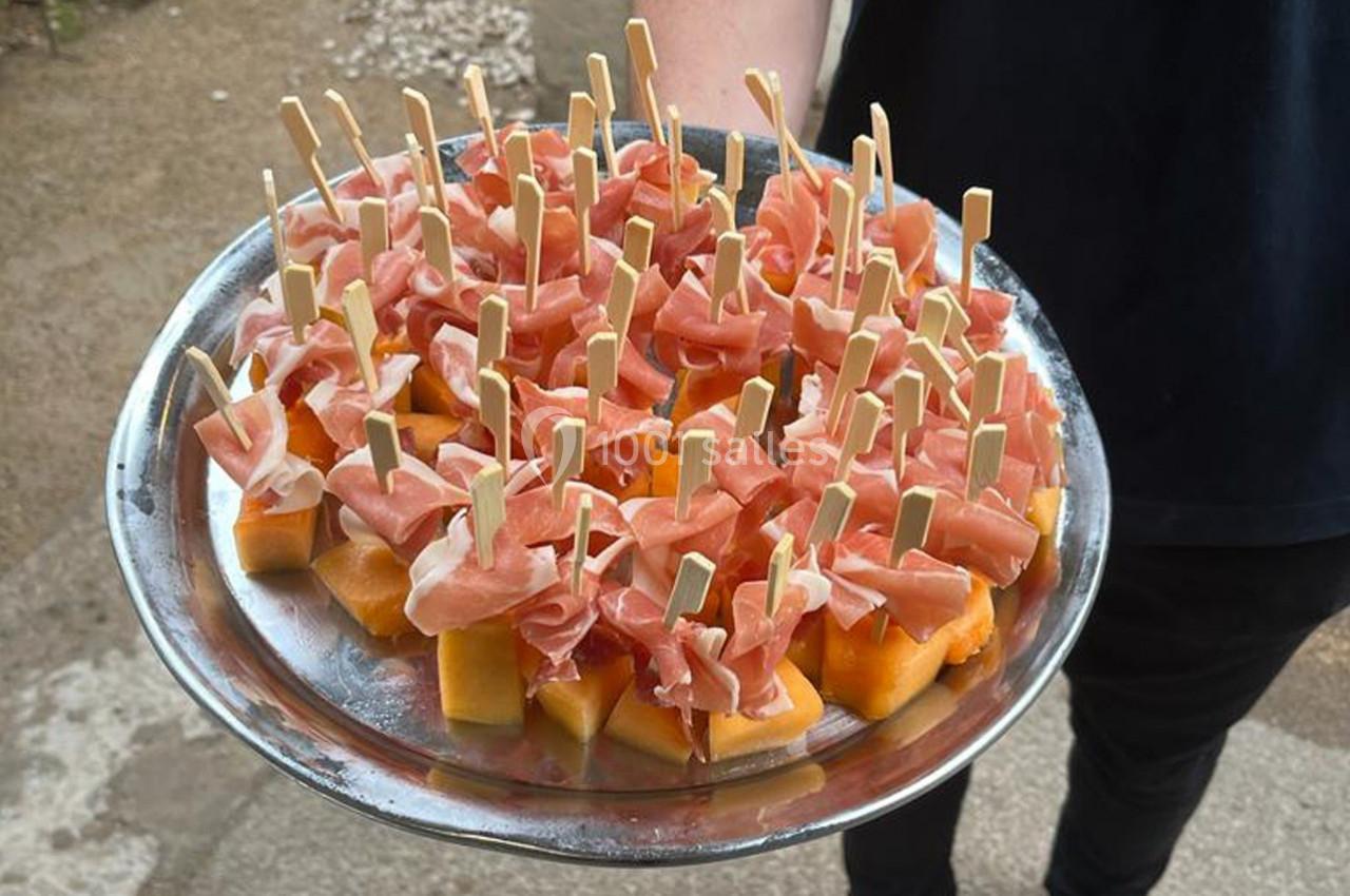 Plateau de brochettes de melon et jambon cru, tenu par une personne en extérieur.