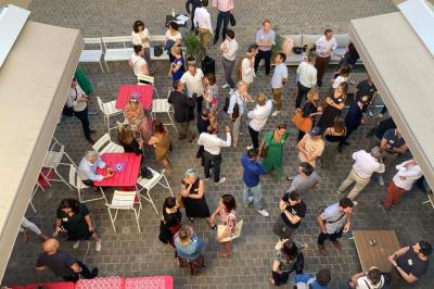 Groupe de personnes rassemblées autour de tables sous des parasols dans une cour extérieure avec végétation.