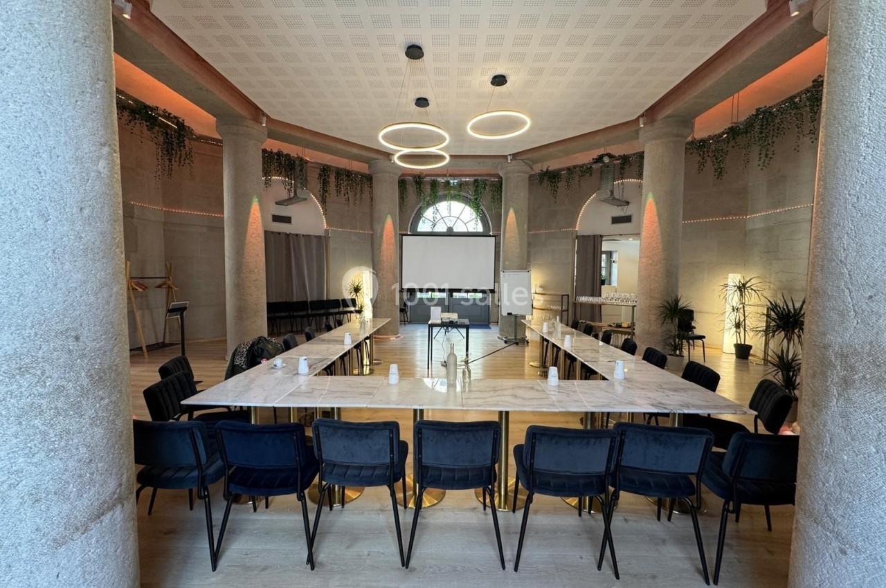 Salle de réunion lumineuse avec une grande table en U, des chaises noires et un écran de projection au fond.