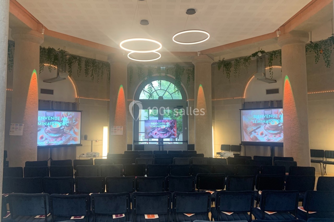 Salle de séminaire lumineuse avec des rangées de chaises noires, deux écrans et une décoration végétale suspendue.