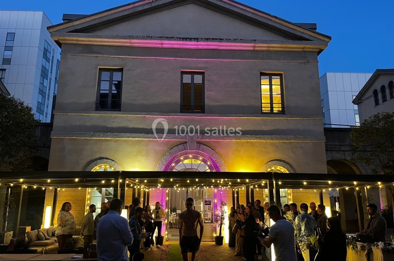Façade éclairée d'un bâtiment ancien avec des invités rassemblés en soirée sous une pergola lumineuse.